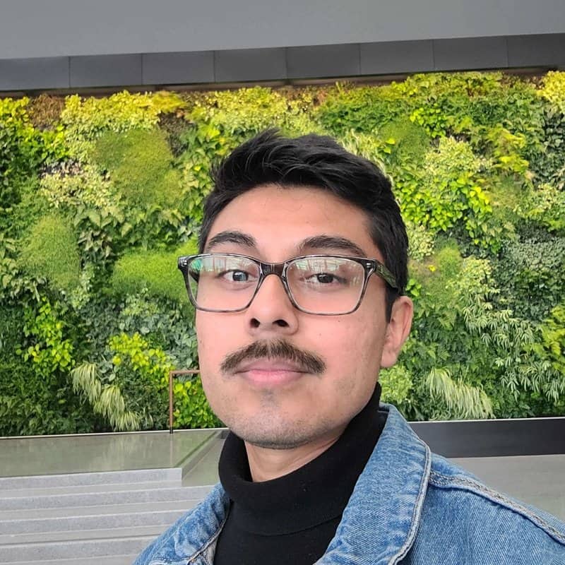 Vibrant green vertical garden wall behind confident young man with glasses and mustache, outdoor modern setting.